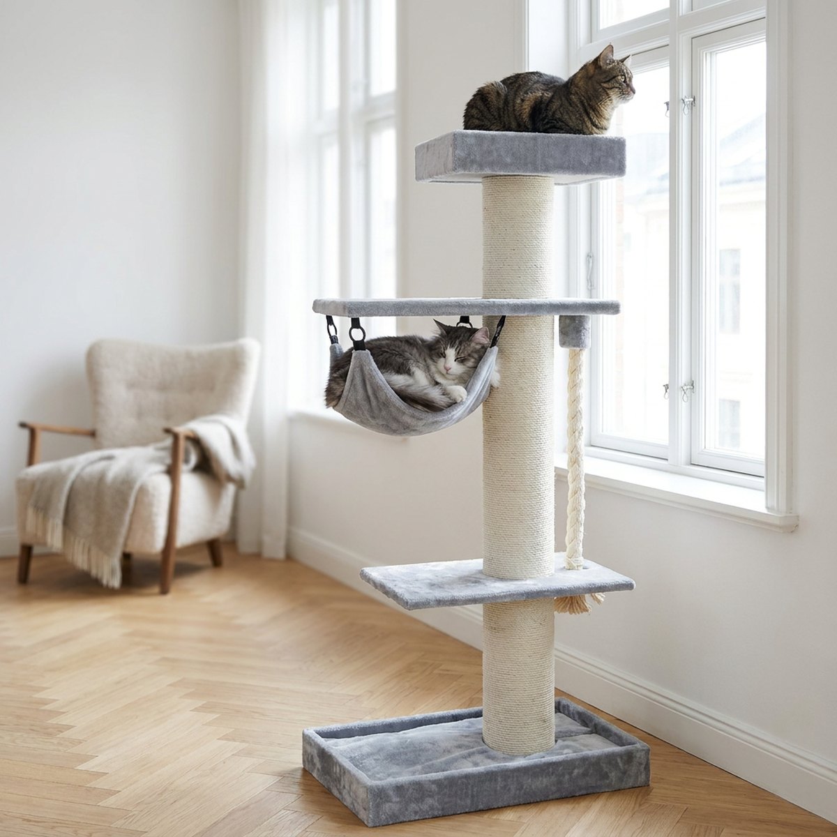 Två katter – en i hängmattan och en på topplattformen – på ett stort grått klösträd med hammock i ett rymligt skandinaviskt rum